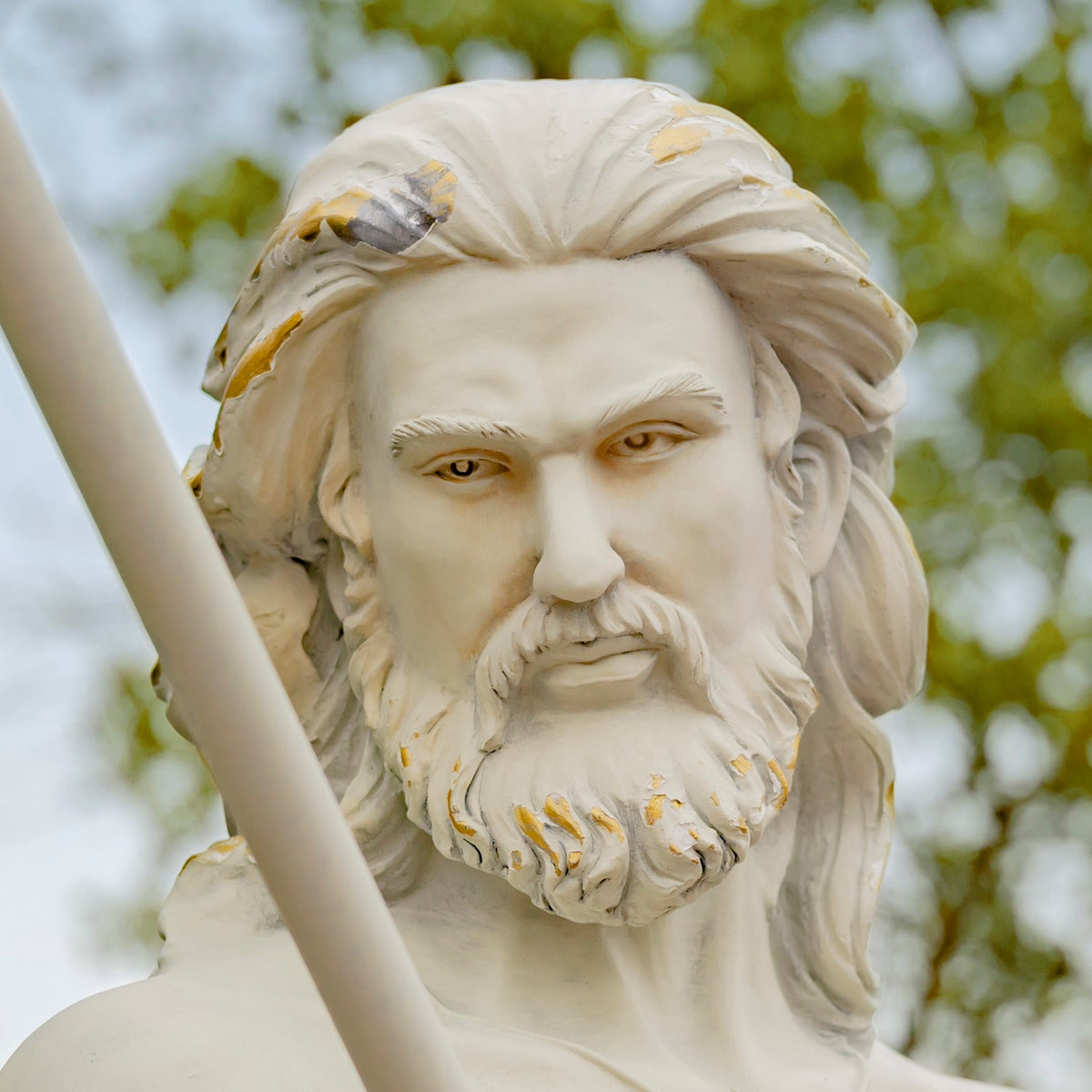 Close-up of a bearded merman statue’s face and flowing hair, with a trident shaft in the foreground.