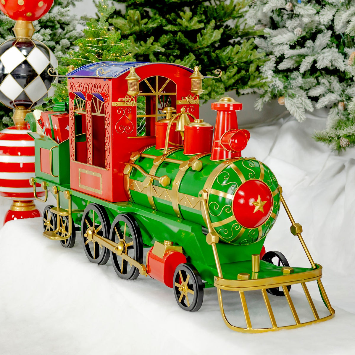Green and red decorative train with a carriage and gold trim displayed beneath a snowy Christmas tree.