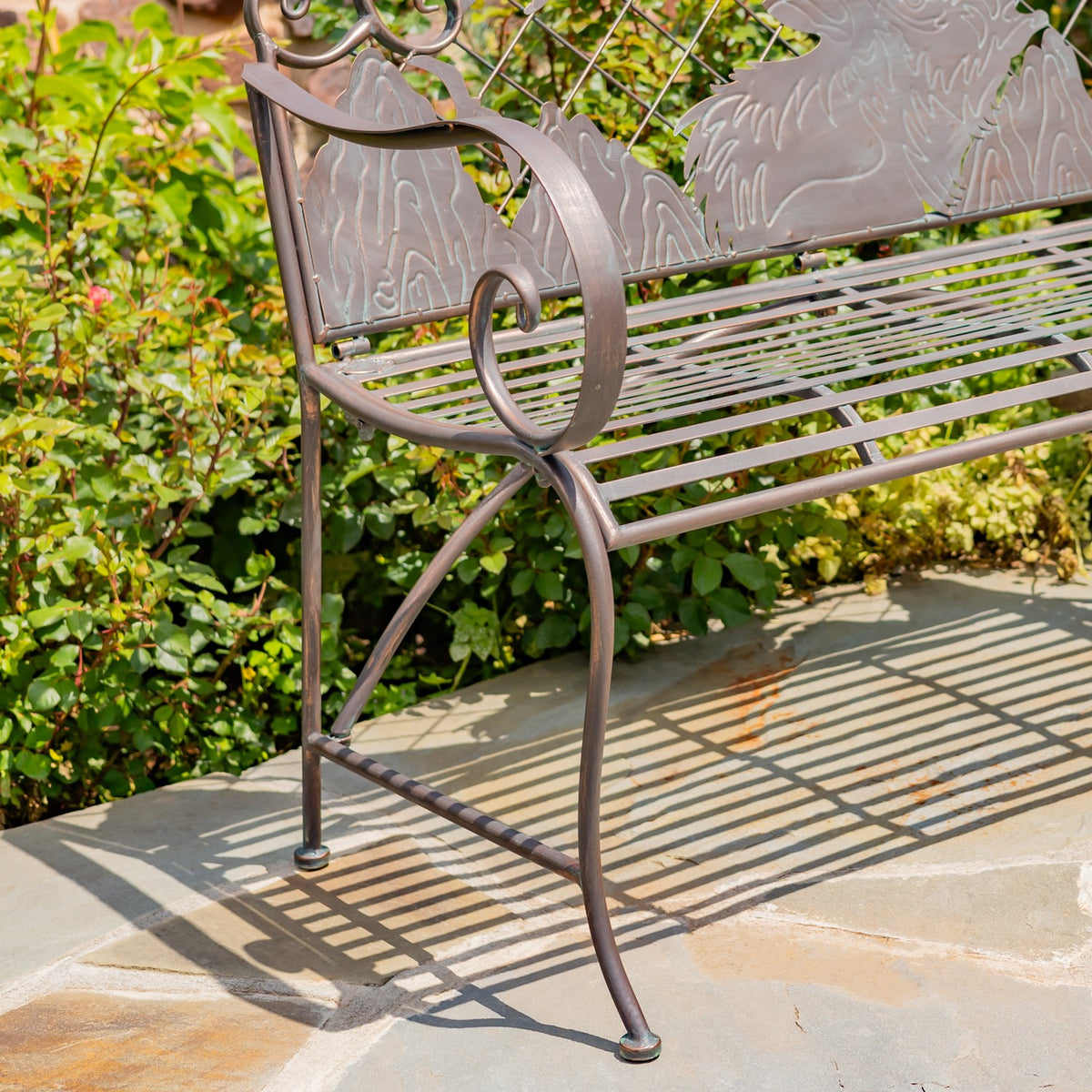 Angled view of an ornate metal garden bench, showing its slatted seat, curved armrest and legs.