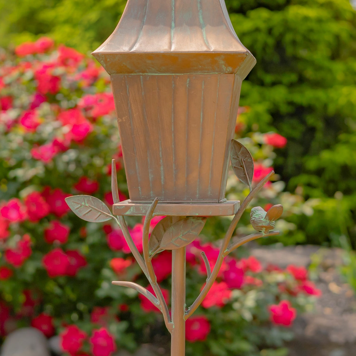 Antique copper birdhouse with raised ribs, leaf-shaped branches and a small bird, among garden flowers.