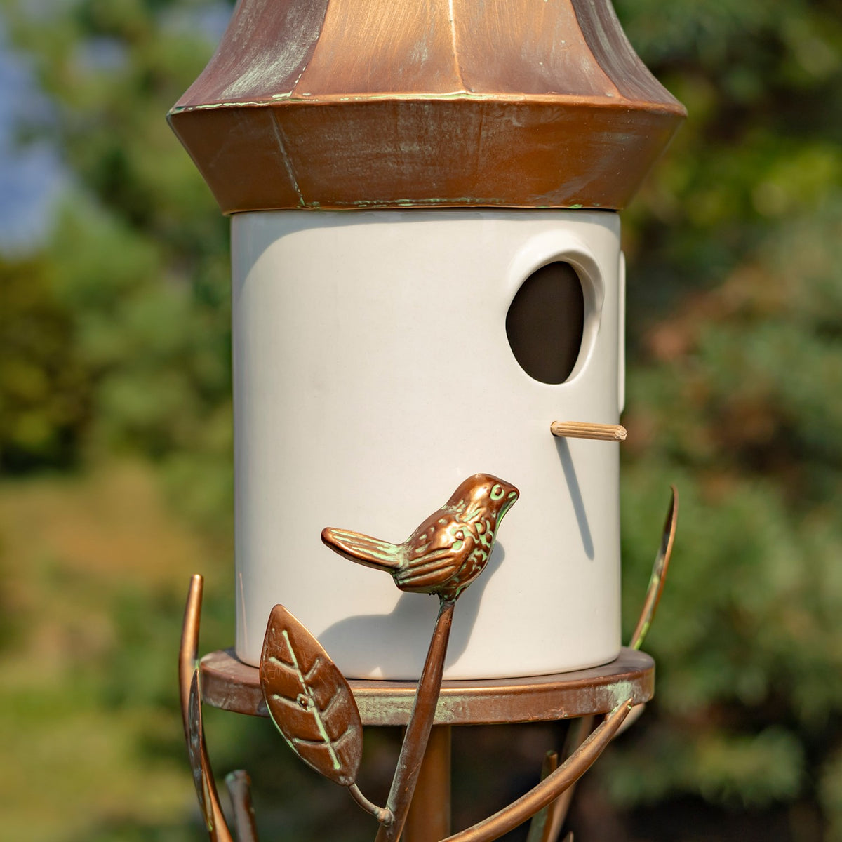 Close-up of a cream birdhouse with a copper-toned roof, bird ornament, and leaf details on its stake.