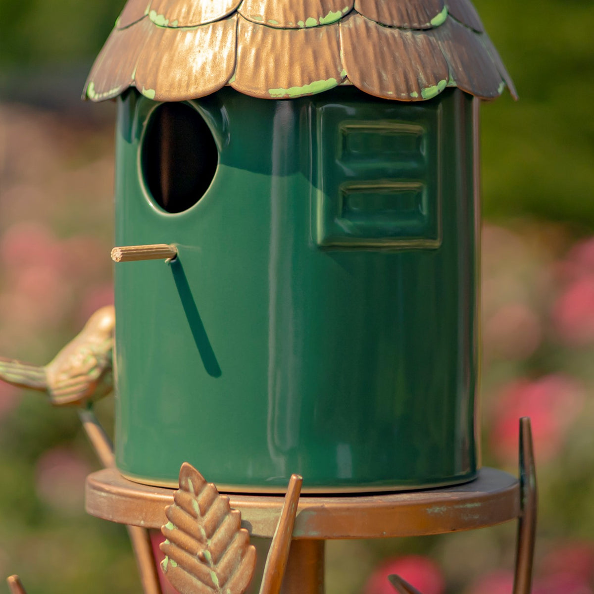 Close-up of a green birdhouse with a round entrance, small perch, and scalloped copper-colored roof.