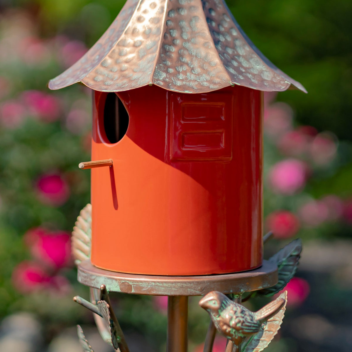 Orange birdhouse with a weathered metal roof, round entrance, and small bird ornament below