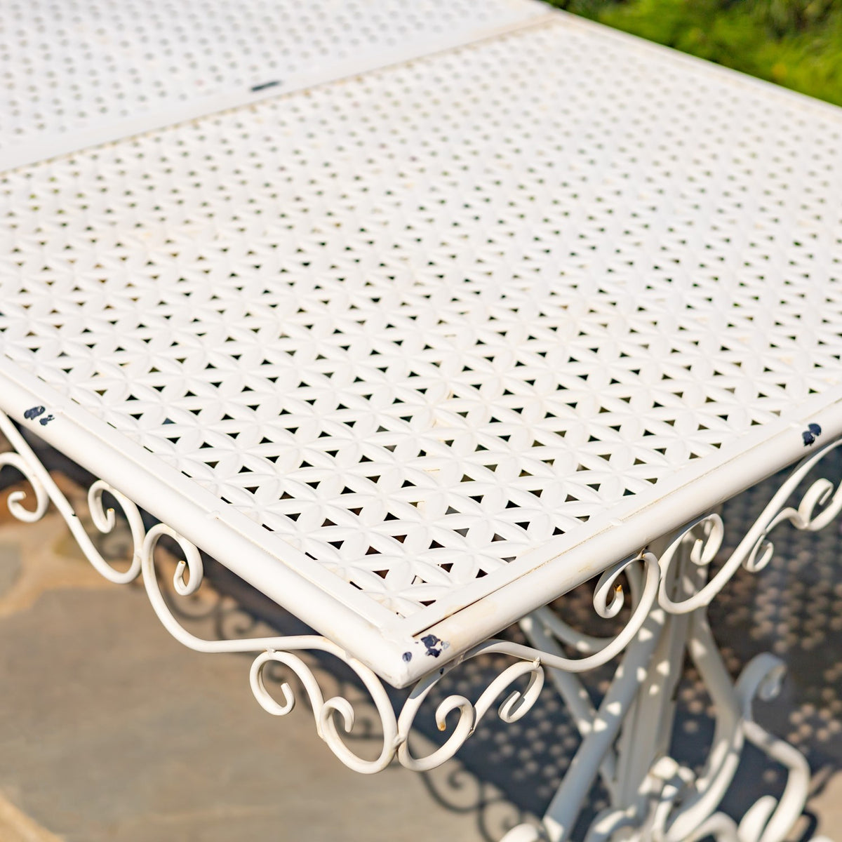 Close-up of a pale metal tabletop with a perforated pattern and curled scrollwork beneath its corner.