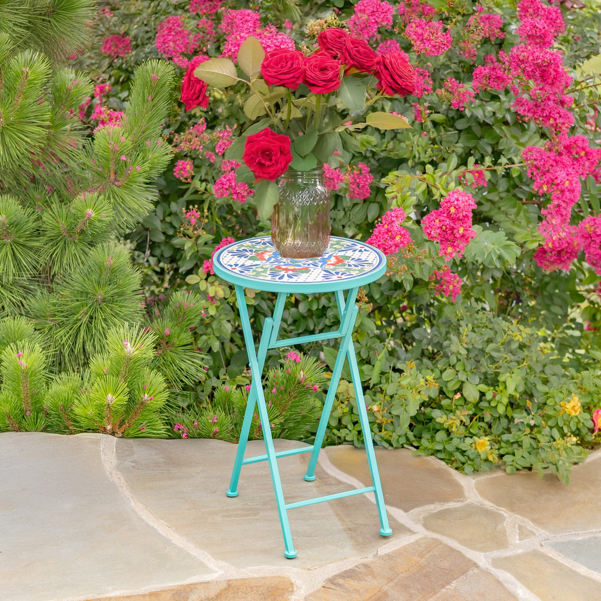 Turquoise folding plant stand with a mosaic top and a vase of red roses in a flower-filled garden.