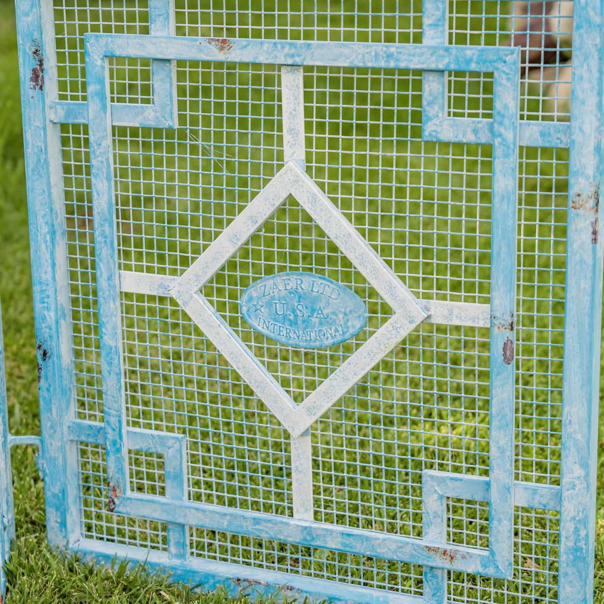 Close-up of a weathered blue and cream metal gate panel with mesh, geometric bars, and an oval center plaque.