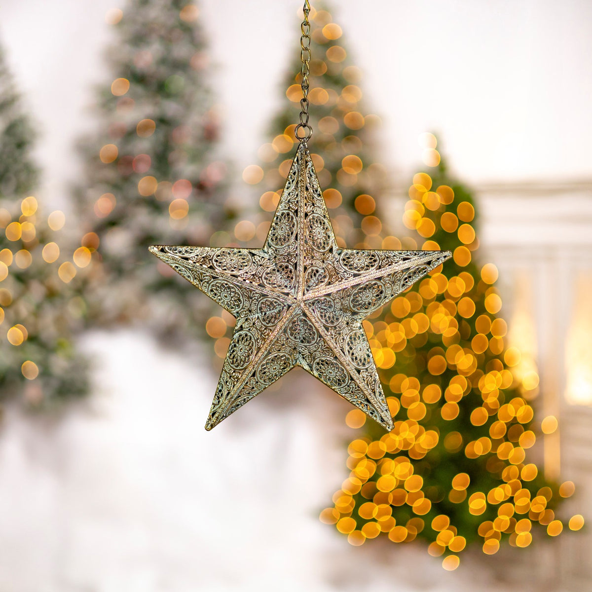 Ornate five-point metal star hanging by a chain in front of softly lit Christmas trees.