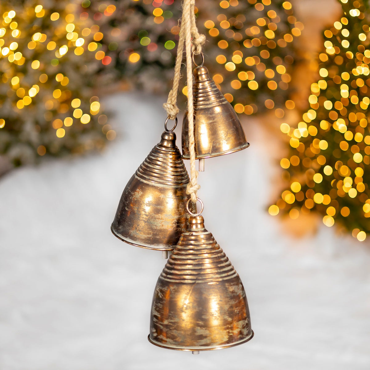 Three antique-gold bells with ridged tops hang at different heights against festive lights.