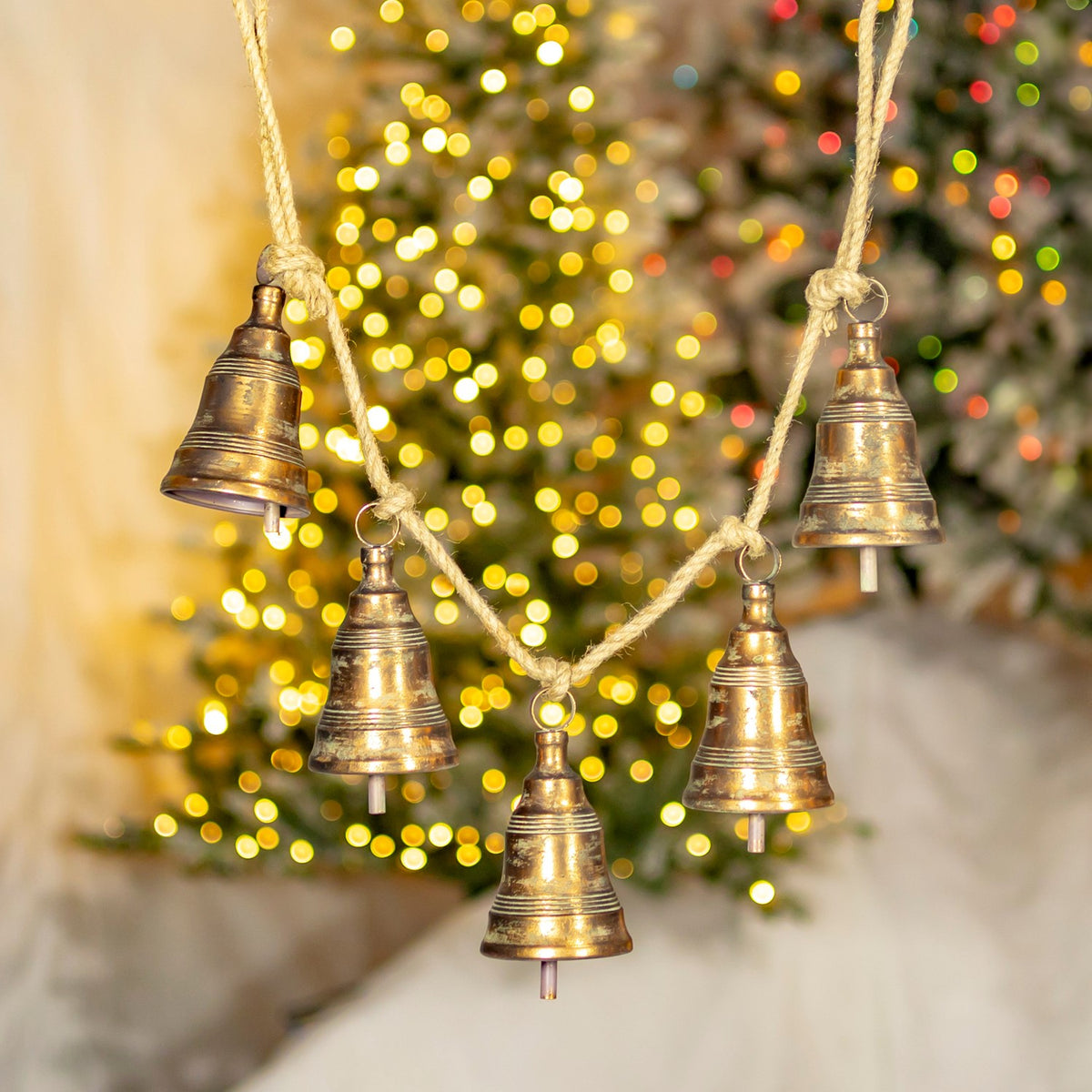 Five antique-gold bells hang from a cord in front of a softly glowing Christmas tree.
