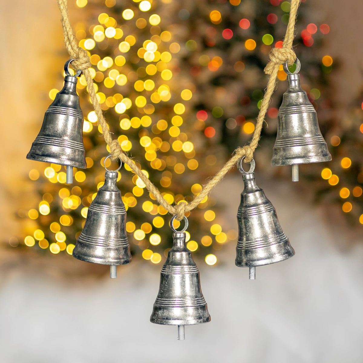 Five antique-silver bells hang from a twine strand against softly glowing Christmas lights.