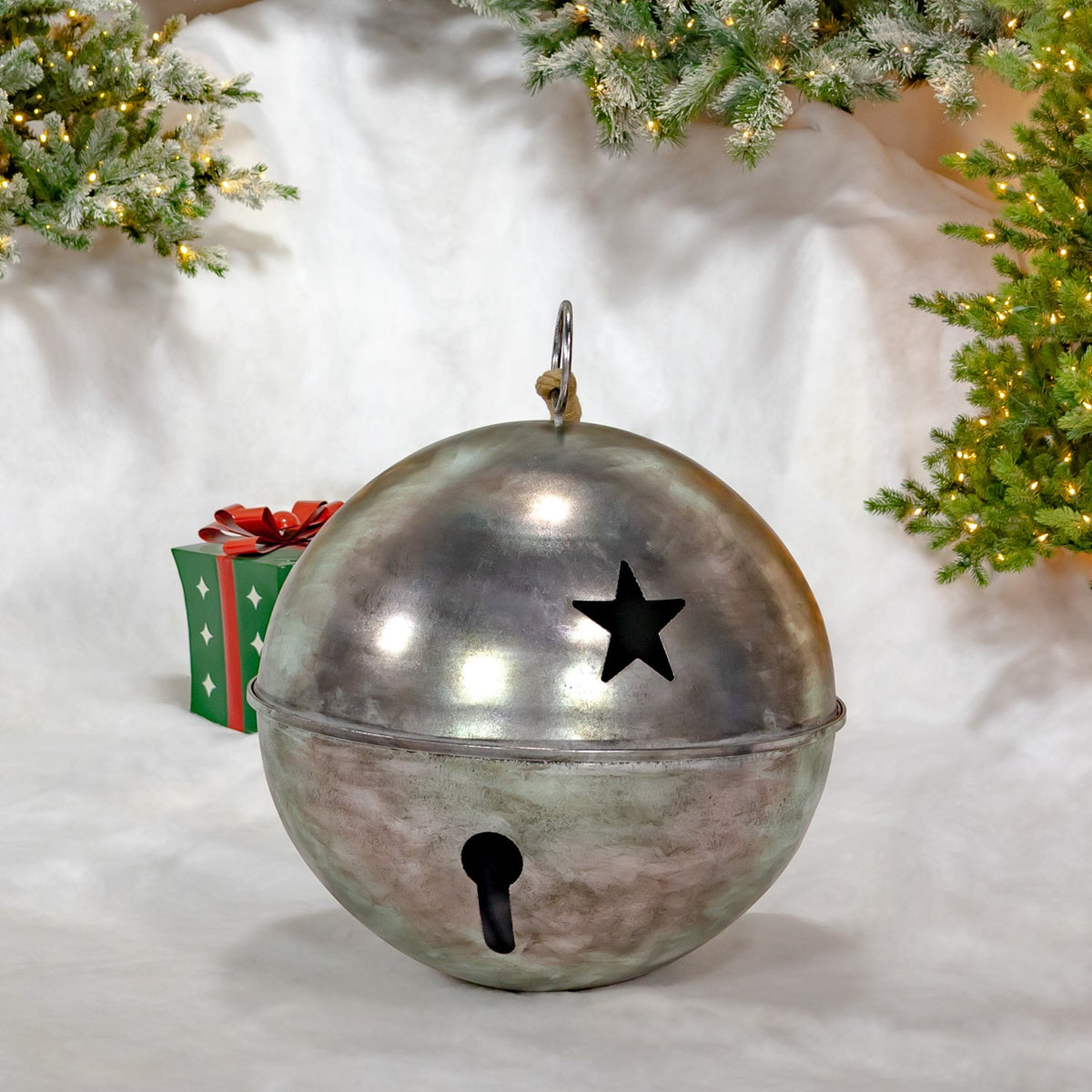 Large silver sleigh bell with a star cutout, displayed on snow beneath lit evergreen branches.