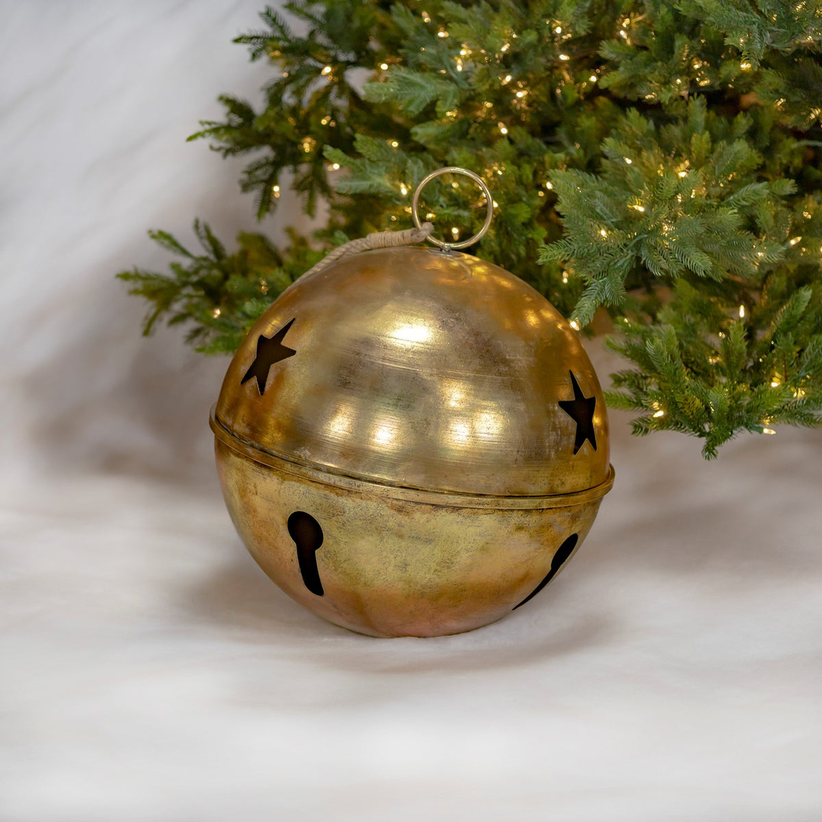 Large antique-gold bell resting beside a lit evergreen tree, with star and keyhole-shaped cutouts.