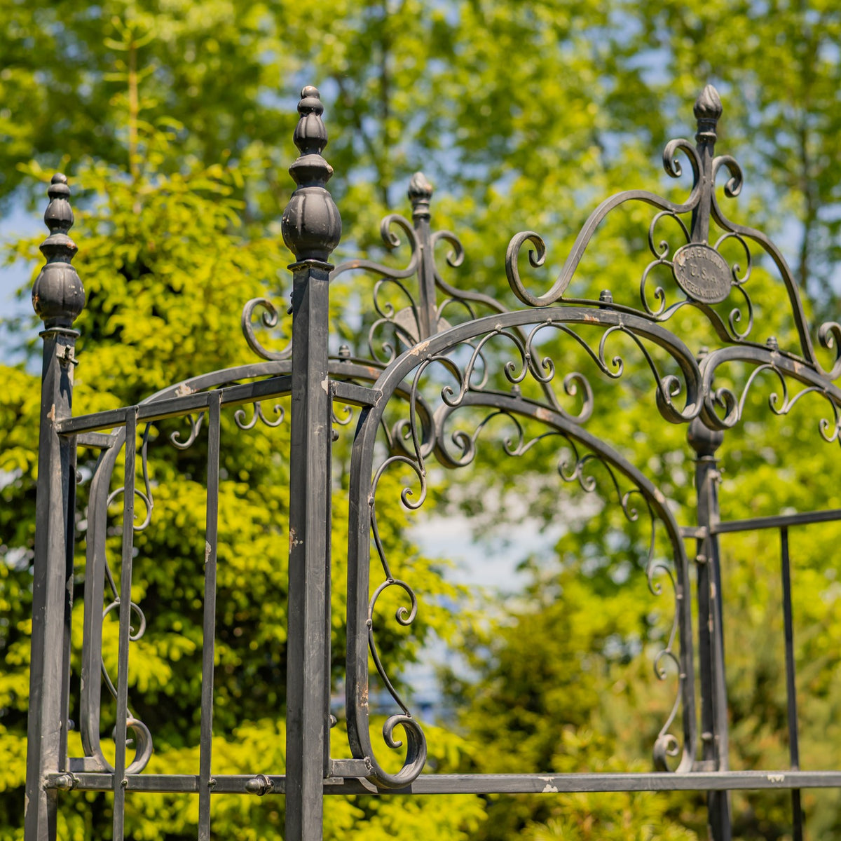 Close view of dark wrought-iron scrollwork and finial-topped posts against leafy trees.