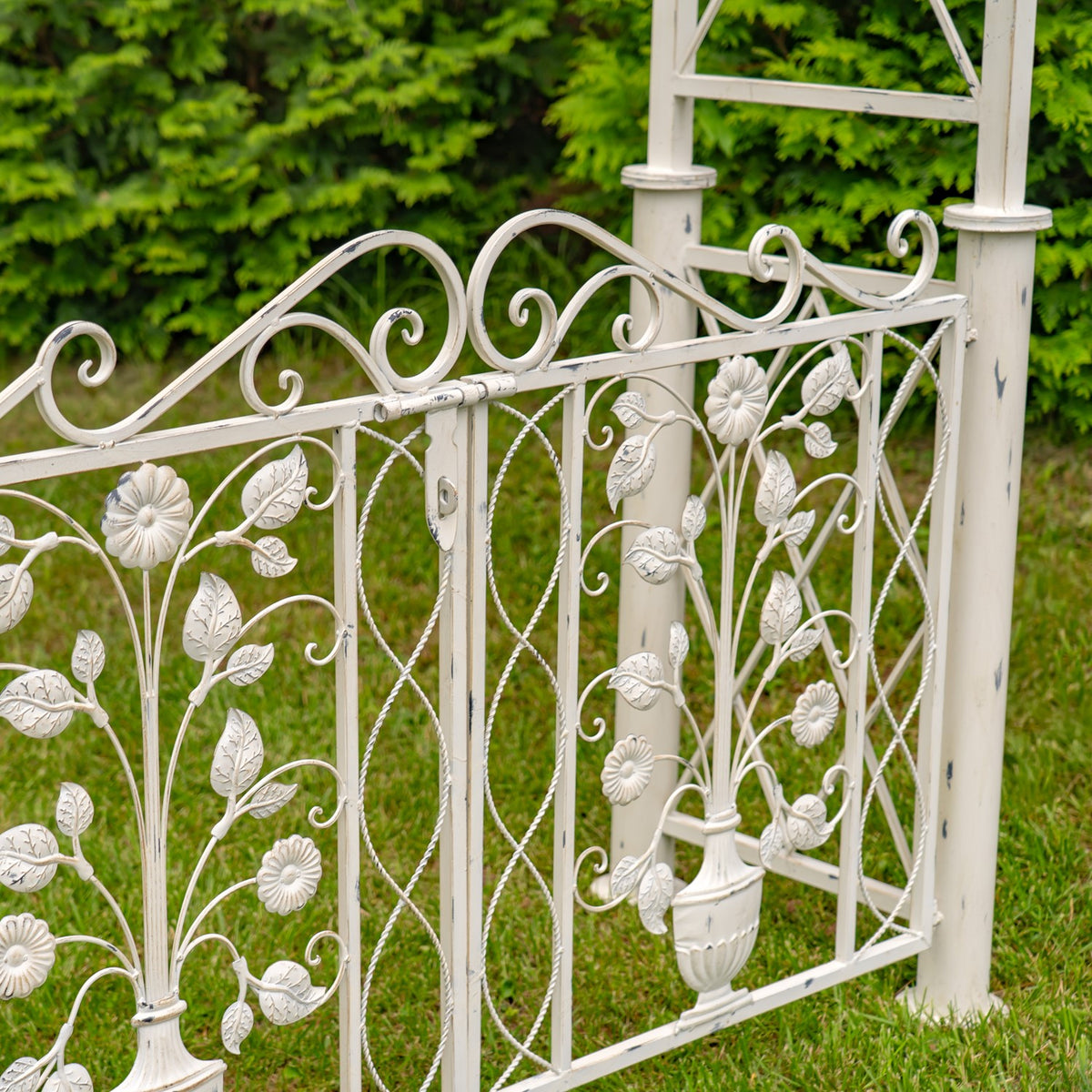 Ornate pale metal gate panel with floral scrollwork between posts in a garden.