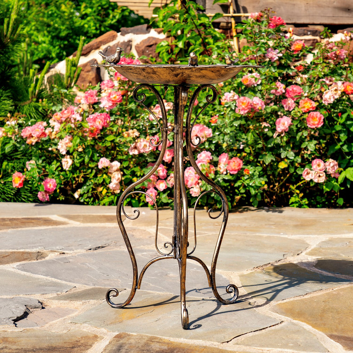 The ornate iron birdbath stands on a stone patio in front of flowering rose bushes.