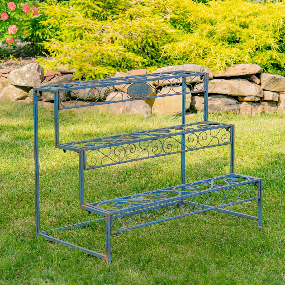 Three-level metal plant stand with scrollwork shelves on grass in a garden.