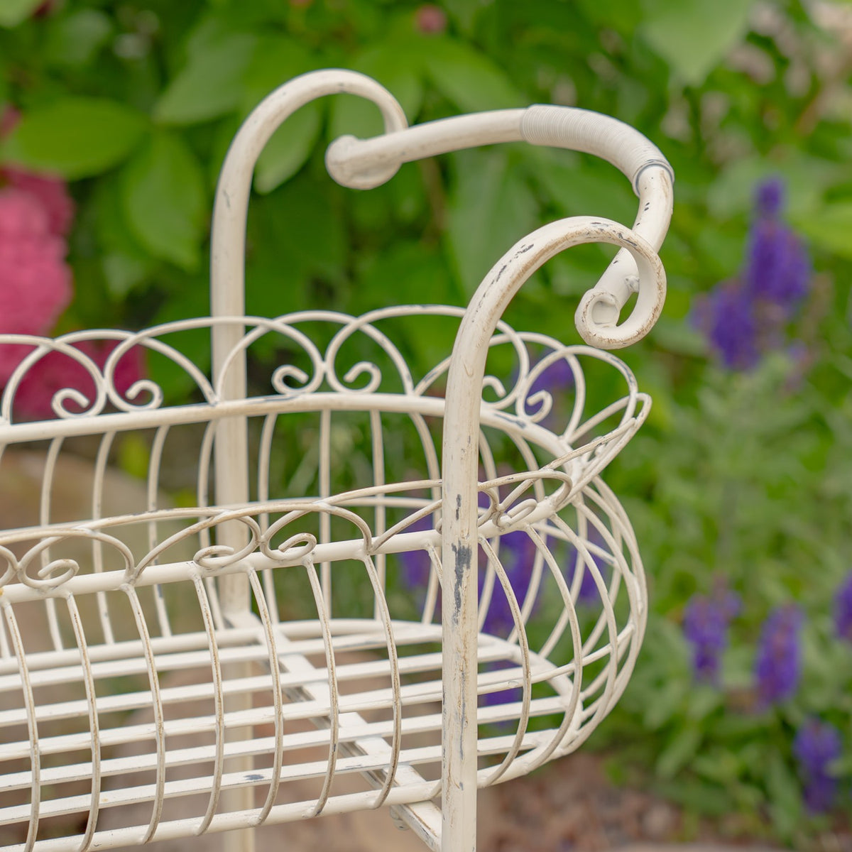 Close-up of the cream metal basket rim, wirework and curled handle against garden greenery.