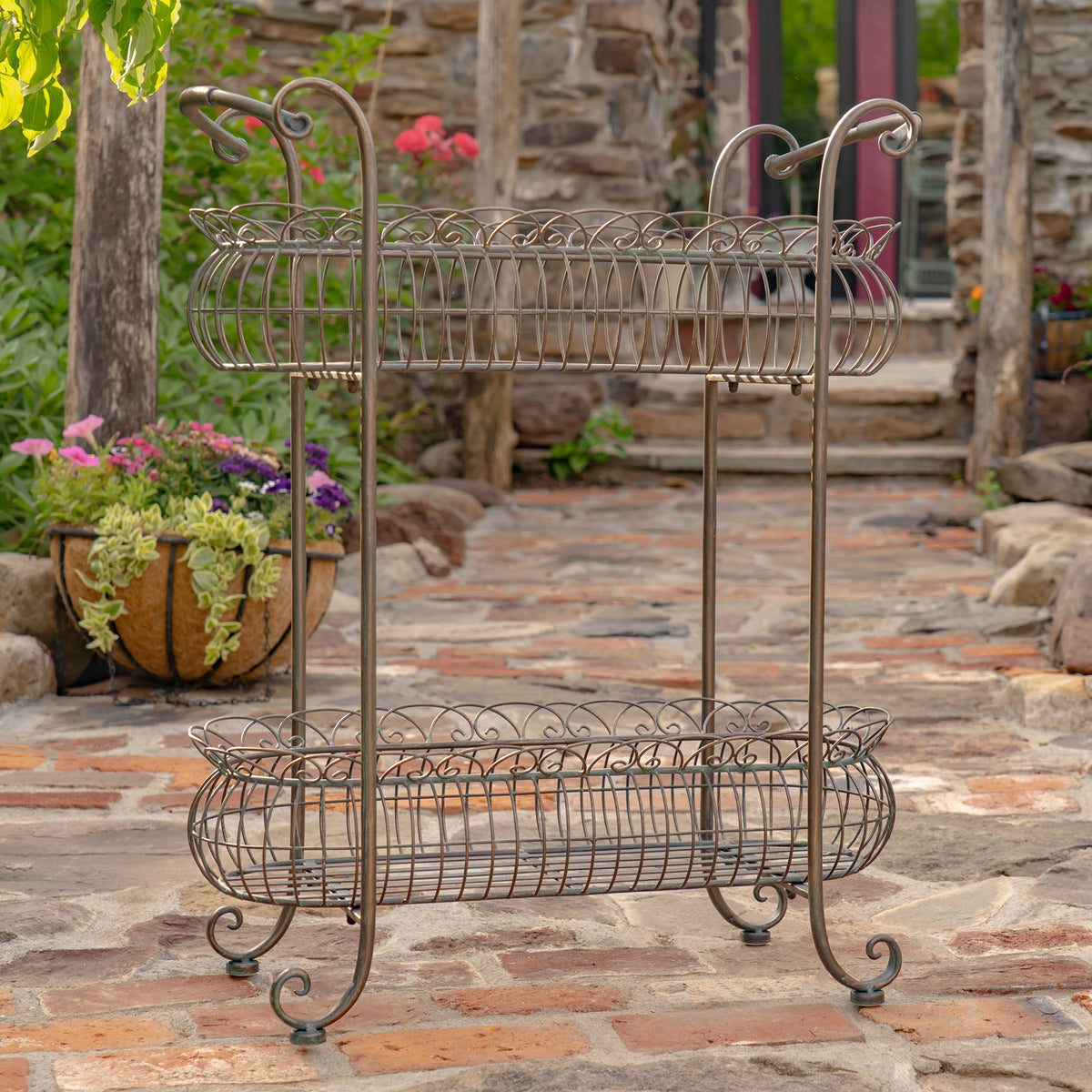 Two-tier oval metal basket stand with curled feet and handles on a stone patio.