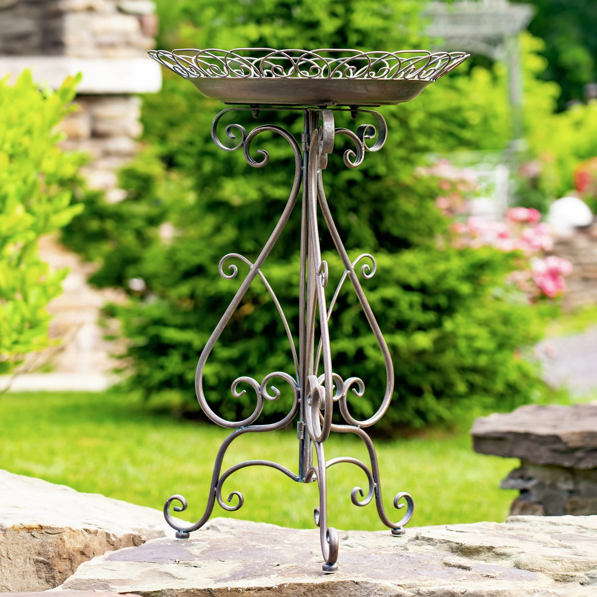 Ornate round metal birdbath on a curled folding stand, displayed on a stone patio among garden greenery.