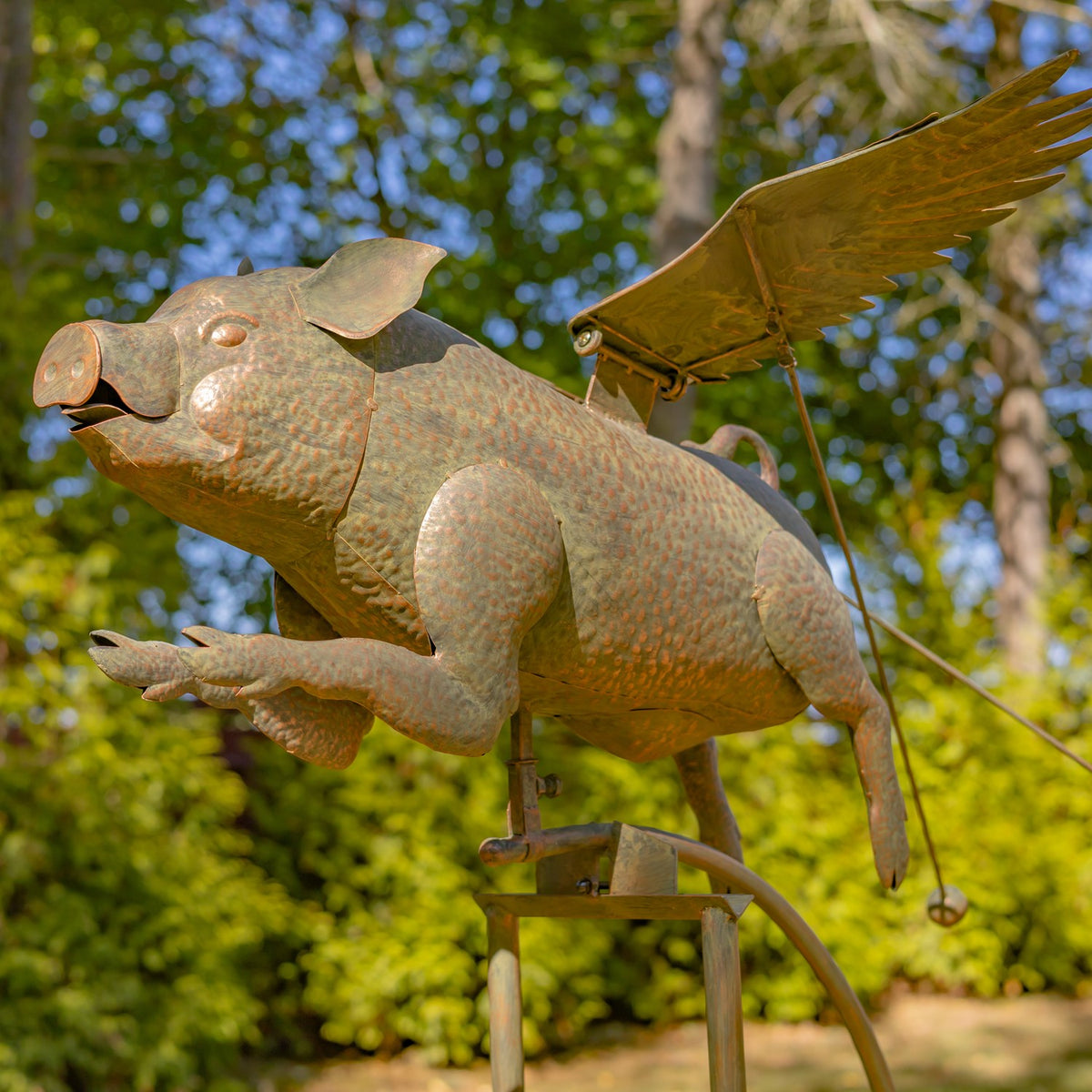 Rust-colored metal pig sculpture with outstretched wings and legs, mounted on a stand against leafy trees.