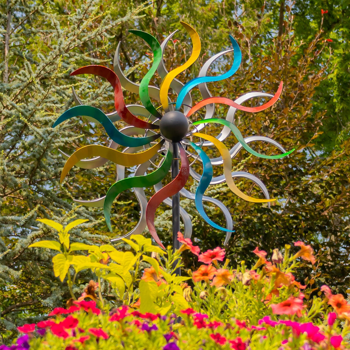 A colorful metal wind spinner rises above bright flowers and leafy garden foliage.