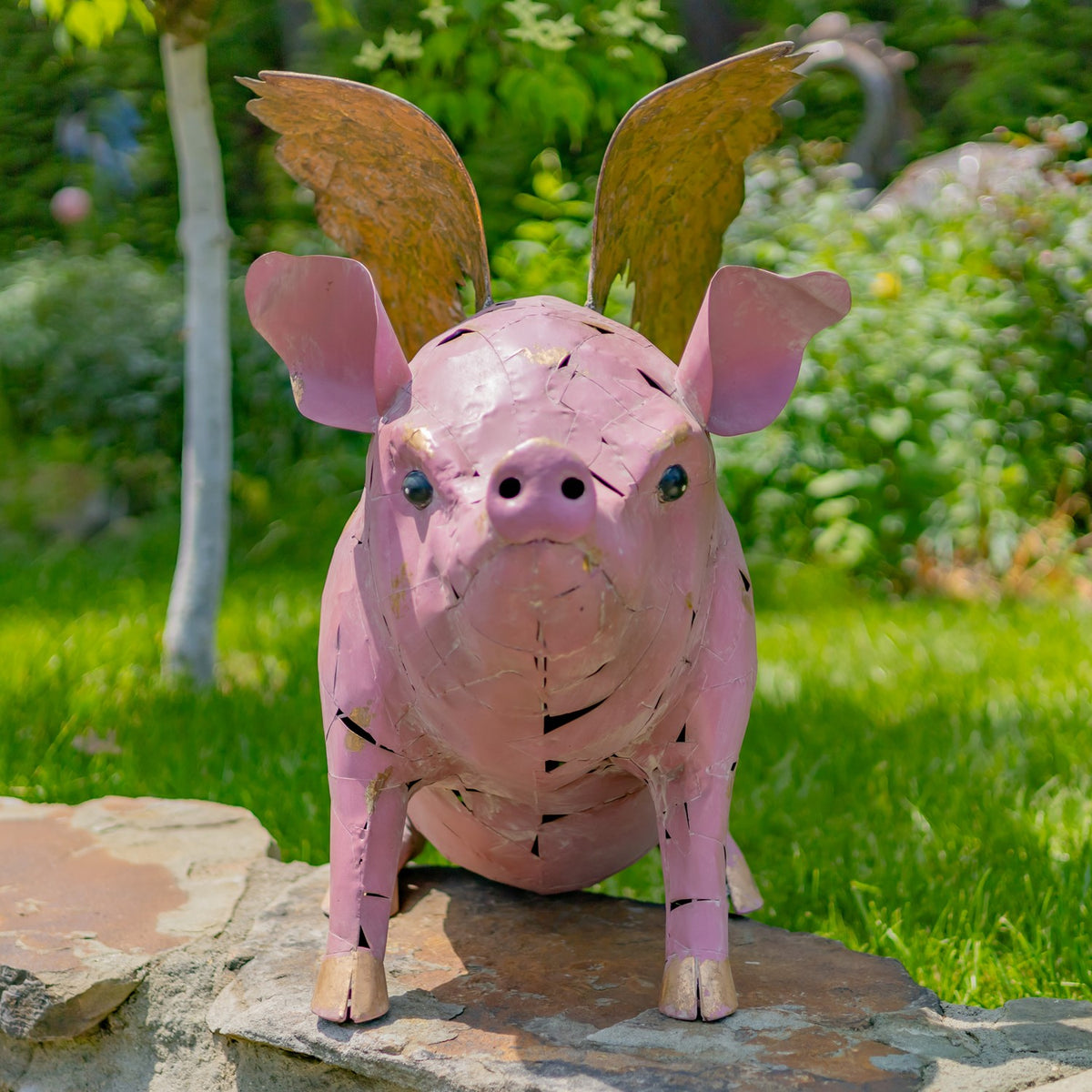 Pink metal pig statue with upright golden wings, standing on a stone surface in a garden.