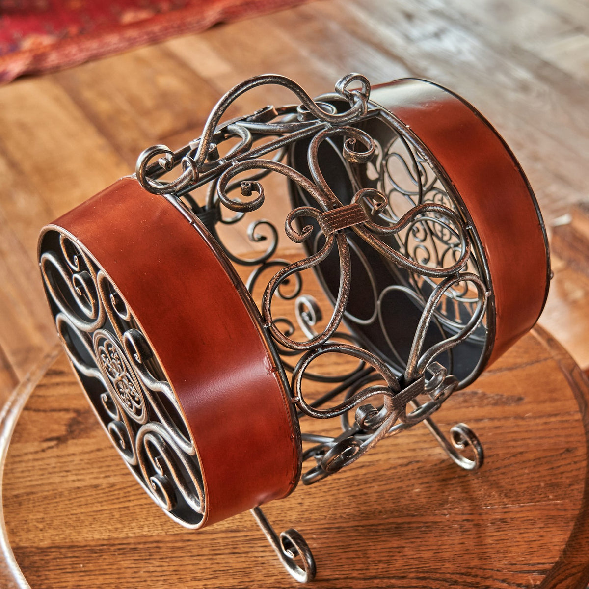 Ornate metal wine rack with reddish-brown bands, curled scrollwork, a top handle, and curved feet.