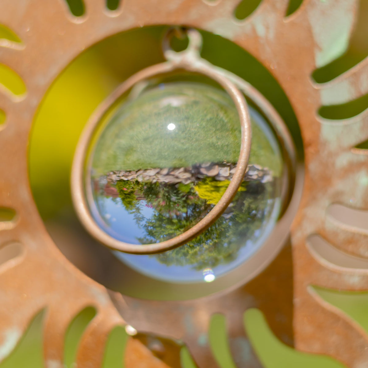 Close-up of a glass orb held in a copper-colored circular frame, reflecting the garden around it.