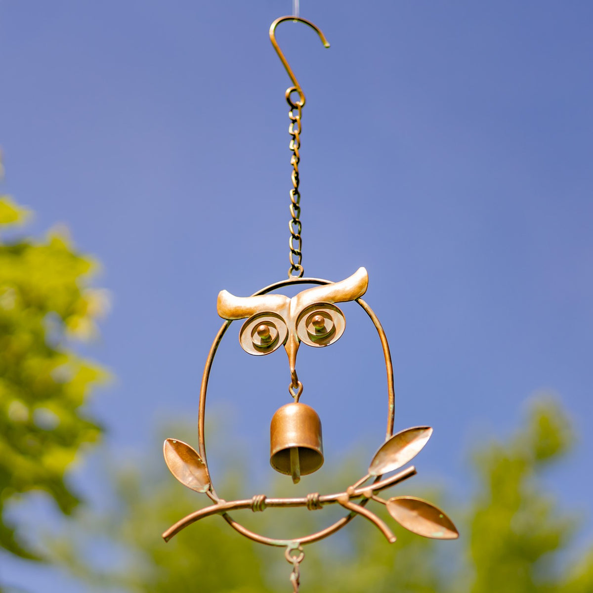 Antique copper owl rain chain hanging against a blue sky, with a small bell and leafy branch details.