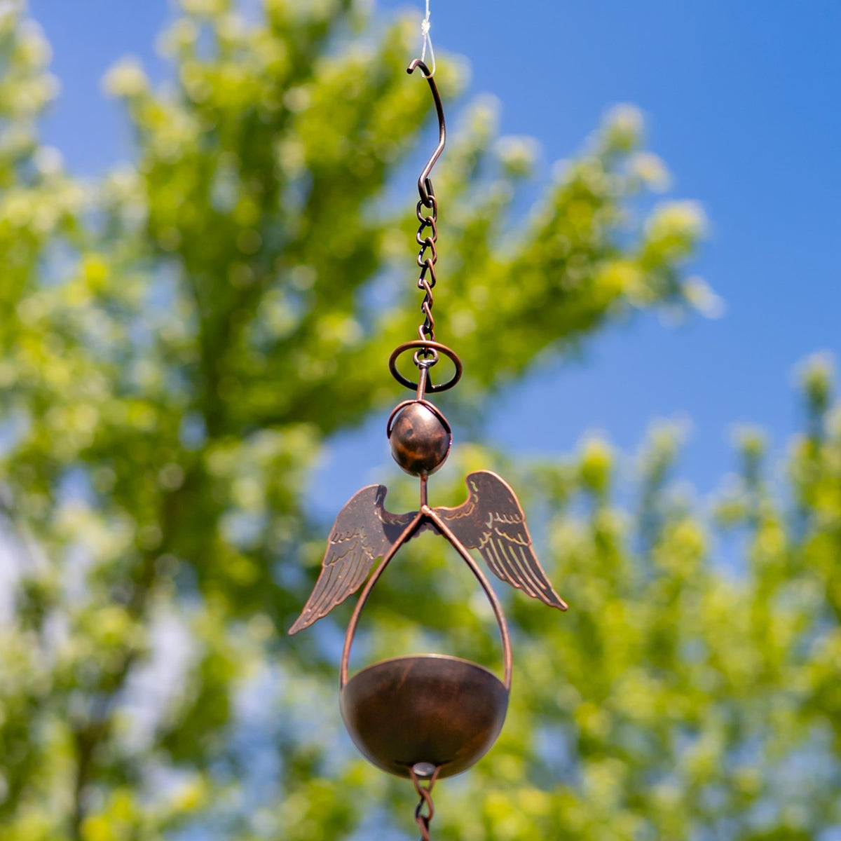 Close-up of a bronze-toned rain chain with an angel figure and bowl-shaped cups against a leafy sky.