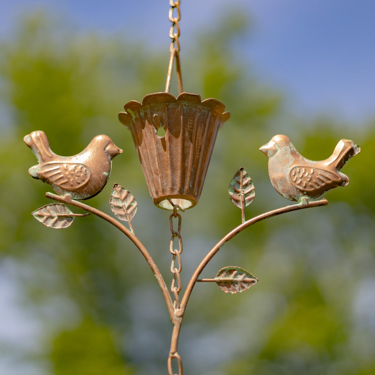 Close view of two metal birds perched on leafy stems around a fluted rain chain cup.