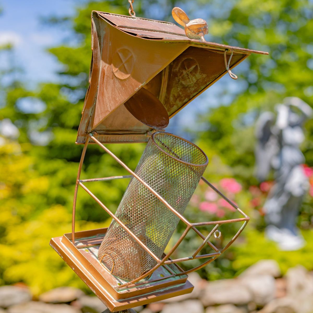 Hanging rust-colored metal birdfeeder with a pagoda roof, tilted mesh seed tube, toadstool accent and small bells.