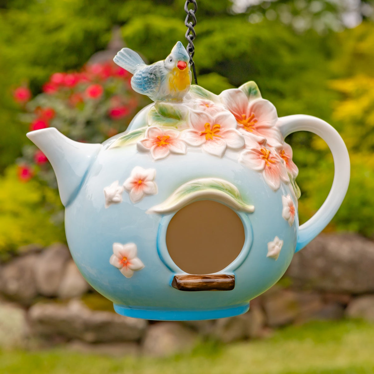 Blue teapot-shaped birdhouse hanging outdoors, decorated with pink flowers and a small bird above its round entrance.