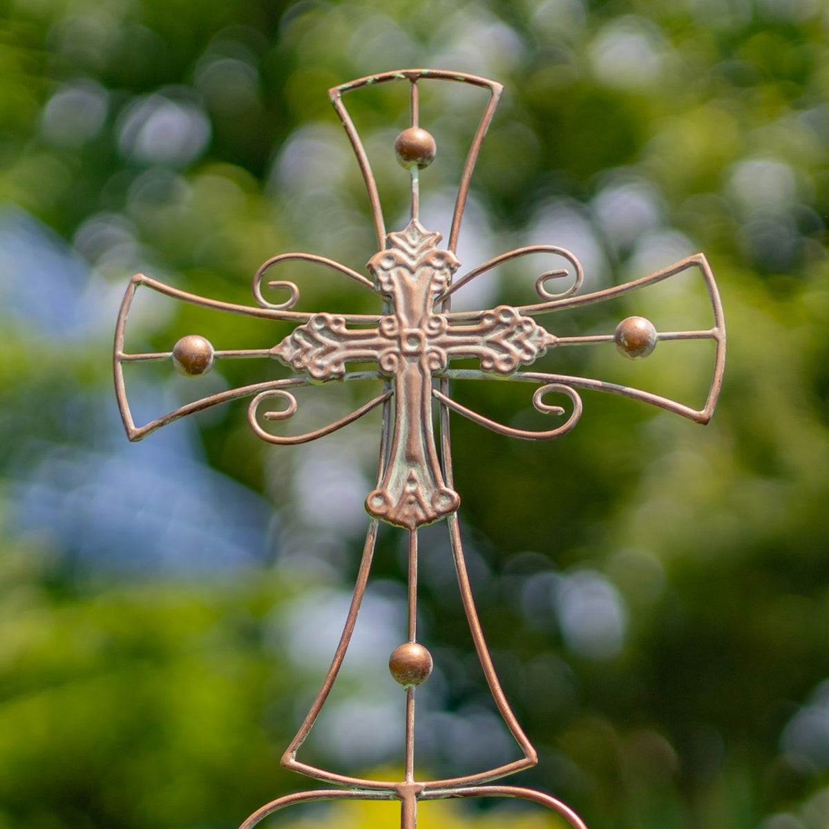Close view of an ornate copper-toned iron cross with curled arms and spherical accents against blurred greenery.