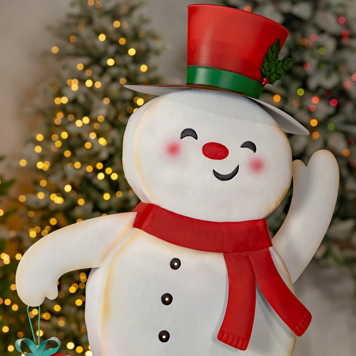Close view of a smiling snowman in a red-and-green hat and scarf, waving in front of a lit tree.