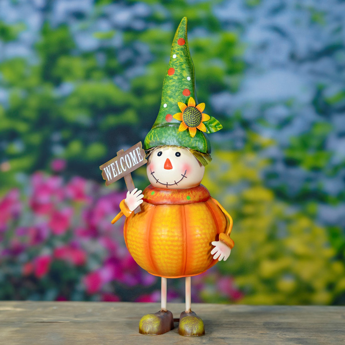 A smiling pumpkin scarecrow with a green sunflower hat holds a small welcome sign outdoors.