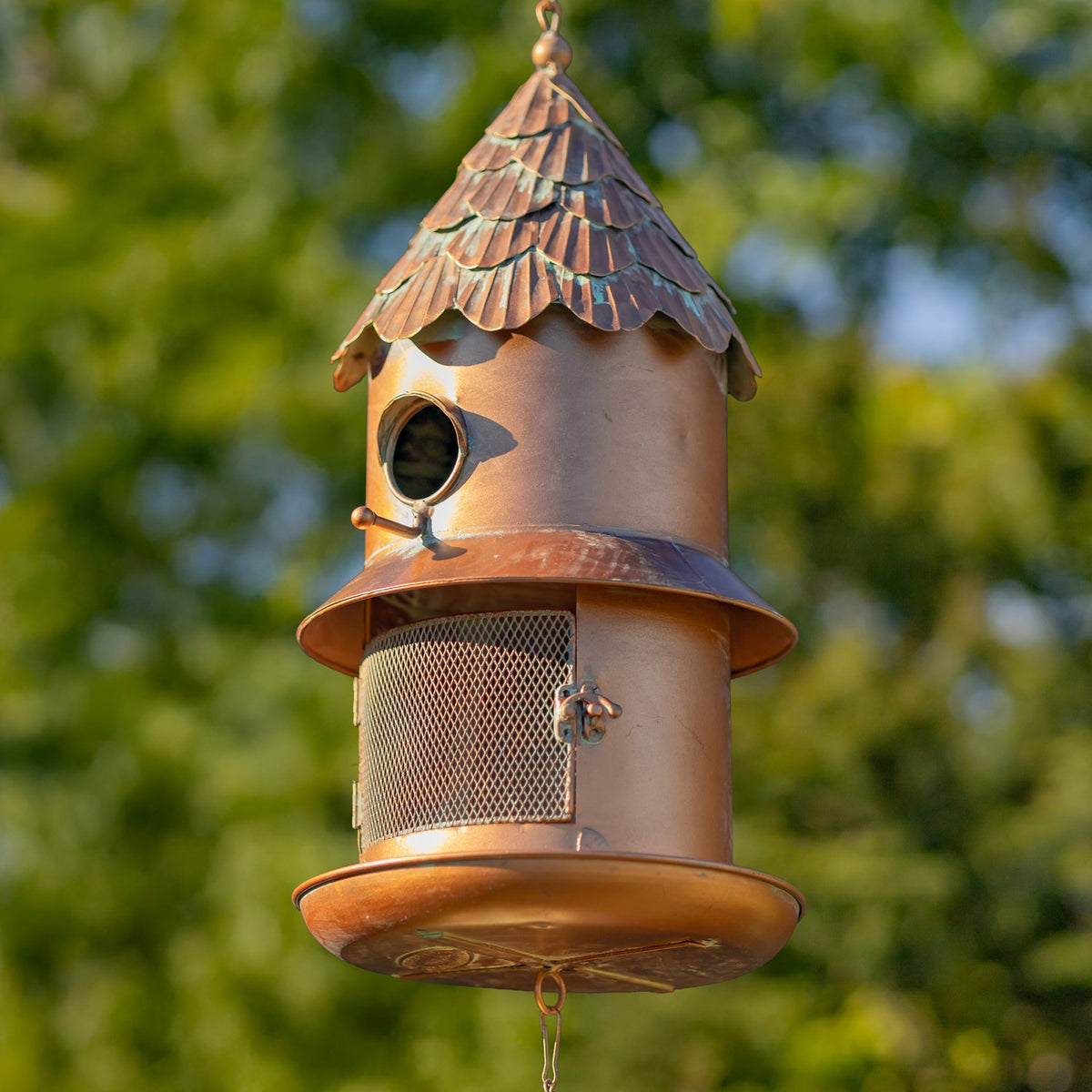 Copper-toned hanging bird feeder with a shingled roof, round entrance, mesh panel and broad tray among trees.