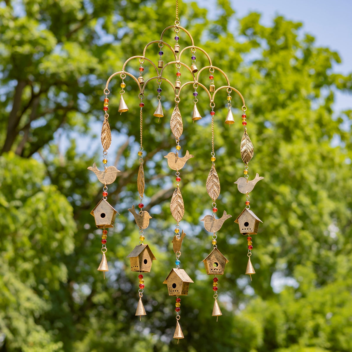 Gold-toned wind chime with dangling bird, leaf, and birdhouse ornaments against leafy trees.