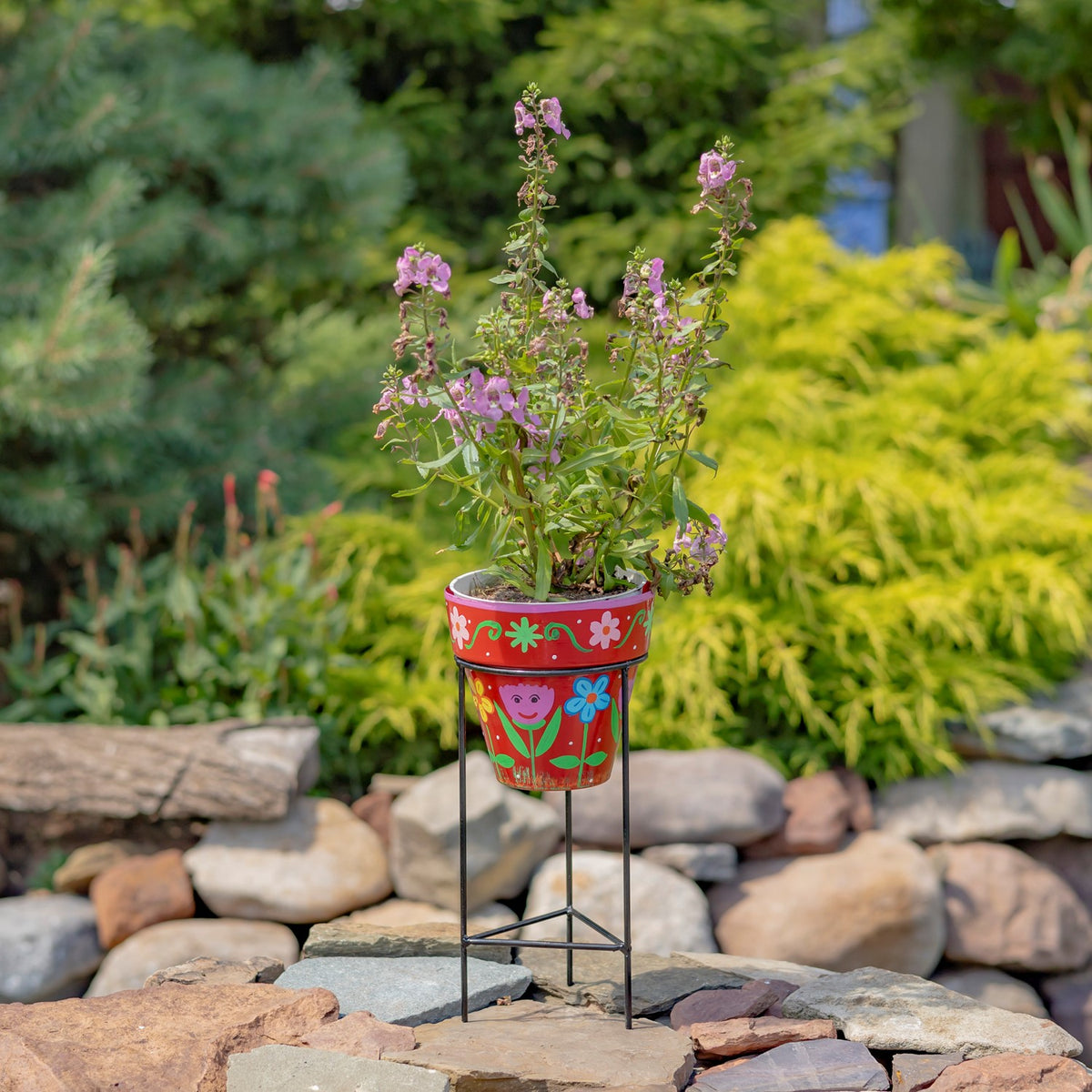 Red hand-painted flowerpot with a pink-flowering plant on a black iron stand among garden stones.