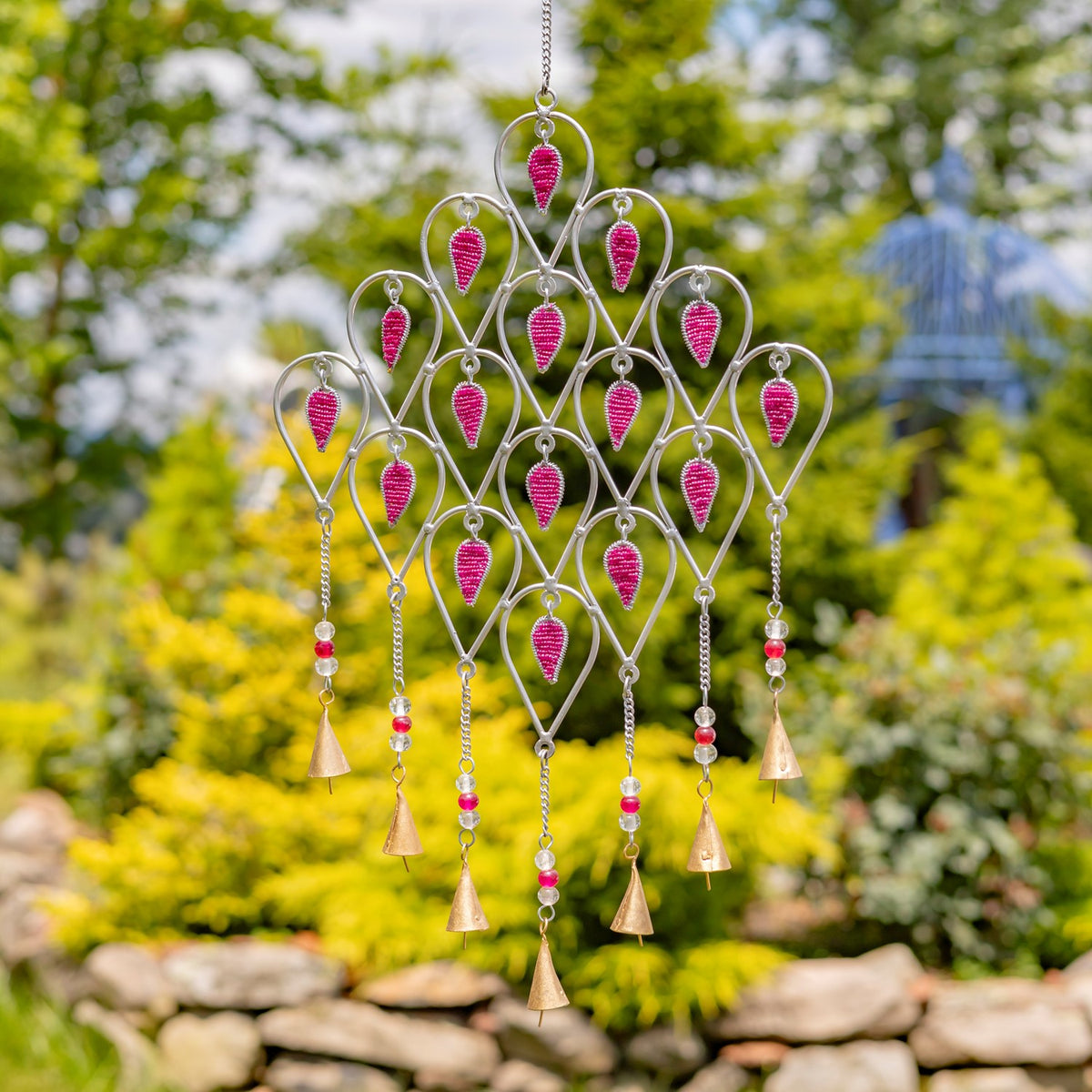 Silver metal wind chime with bright pink accents and dangling beads and bells against a leafy garden.