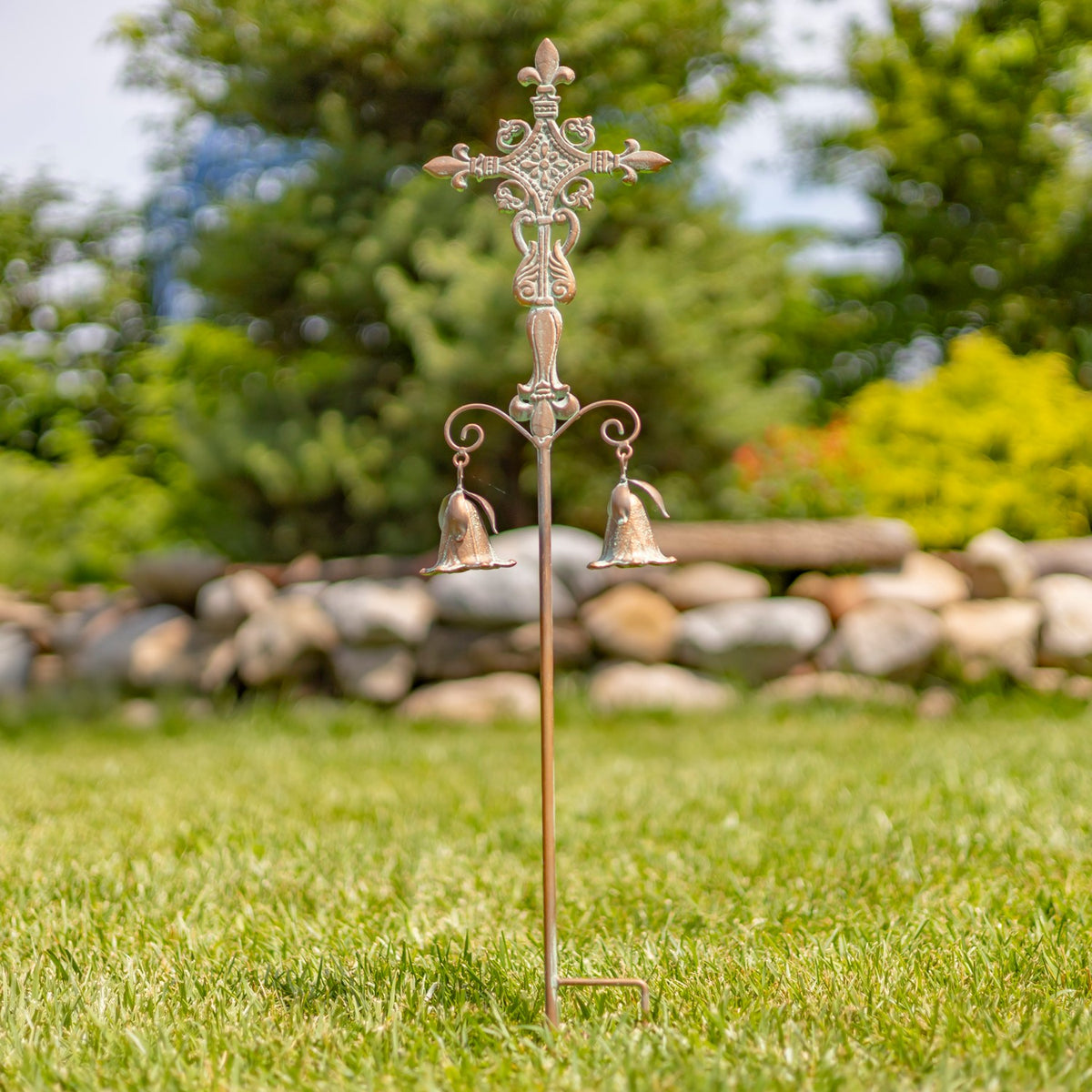 Ornate metal cross garden stake with two hanging bells standing in a grassy garden.