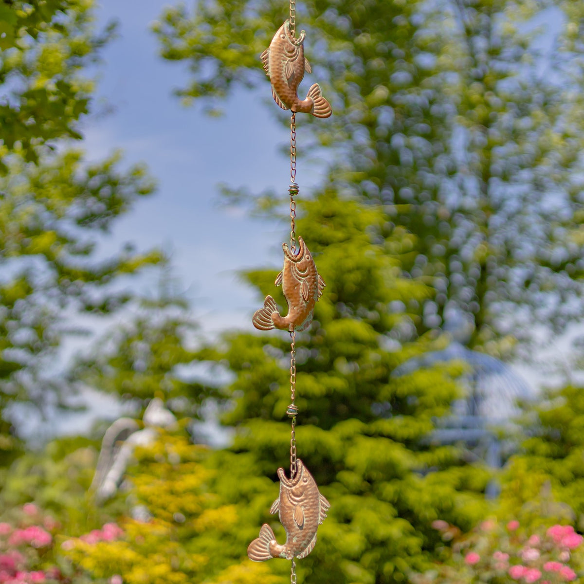 A vertical rain chain with three fish-shaped links hangs against leafy garden greenery.
