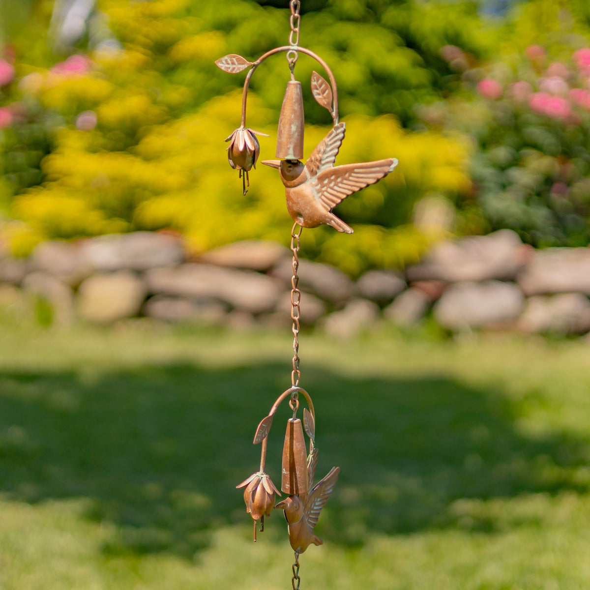 Copper-toned rain chain hangs in a garden, with hummingbird and flower-shaped sections linked vertically.