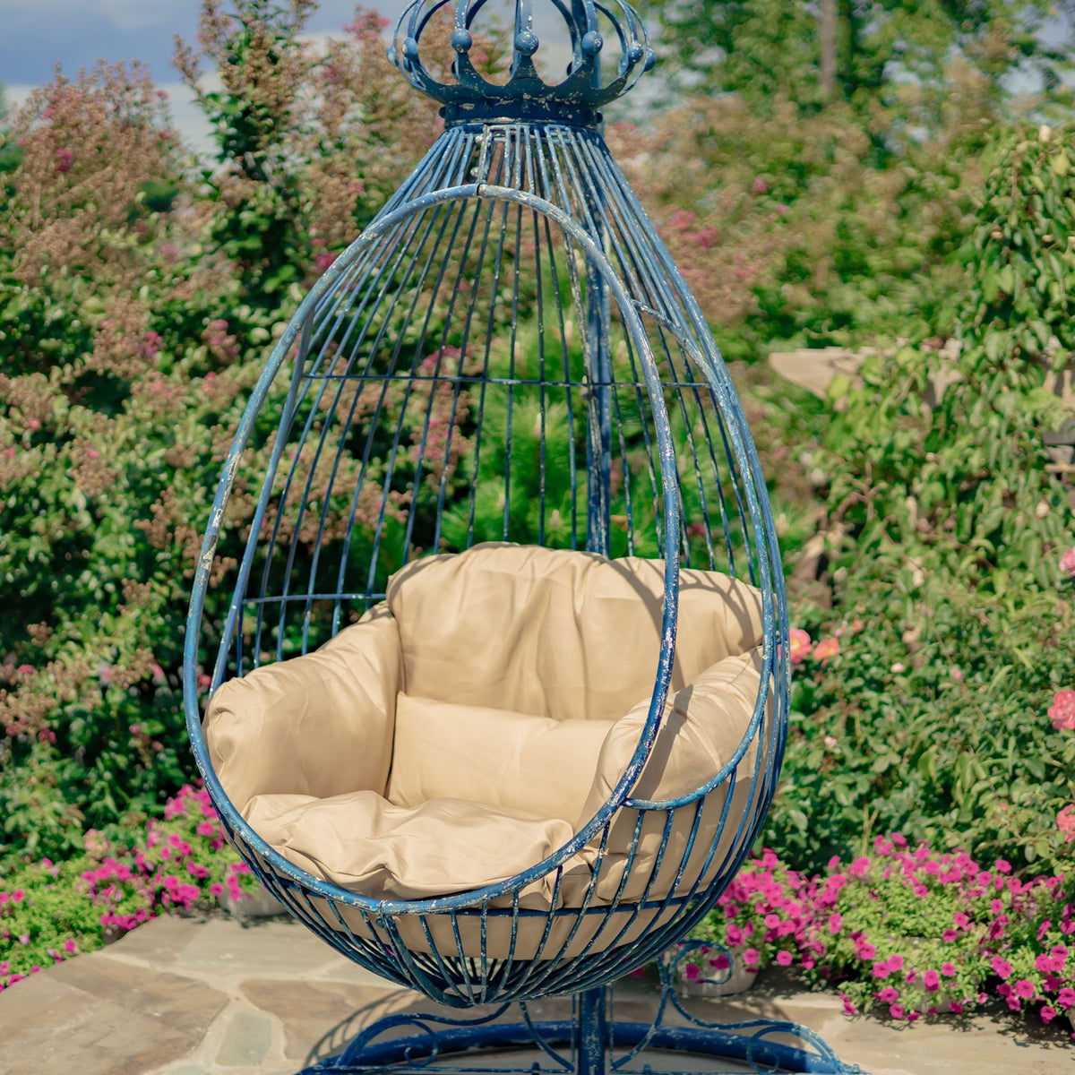 Blue metal egg chair with beige cushions hanging from its stand in a garden.