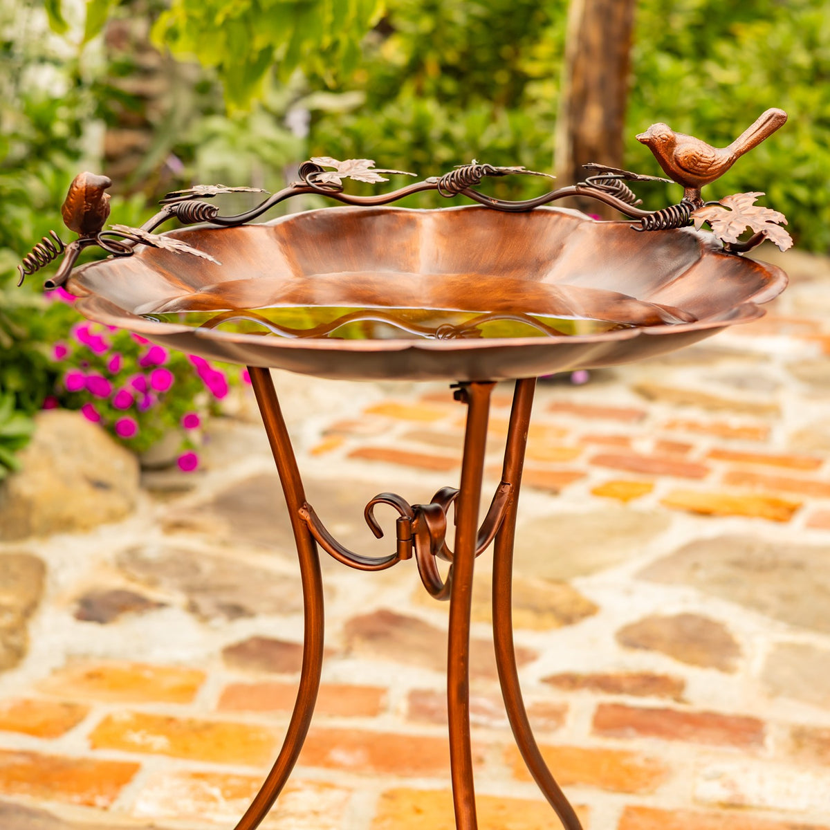 Flower-shaped birdbath with two bird ornaments on a bronze-colored stand, shown on a stone patio.