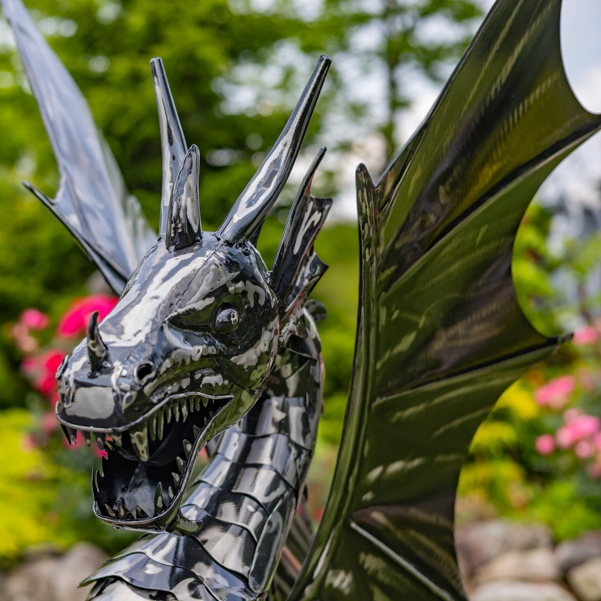 Close-up of a shiny metal dragon’s open mouth, spiked head and large outstretched wing among garden flowers.