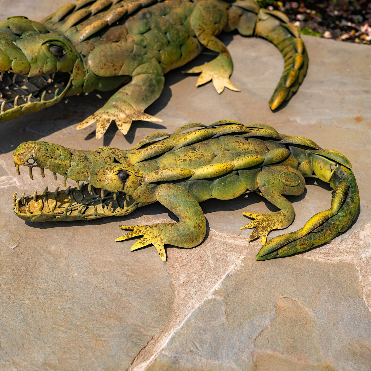 Two mottled green and yellow metal alligator garden decorations lie on a stone surface.