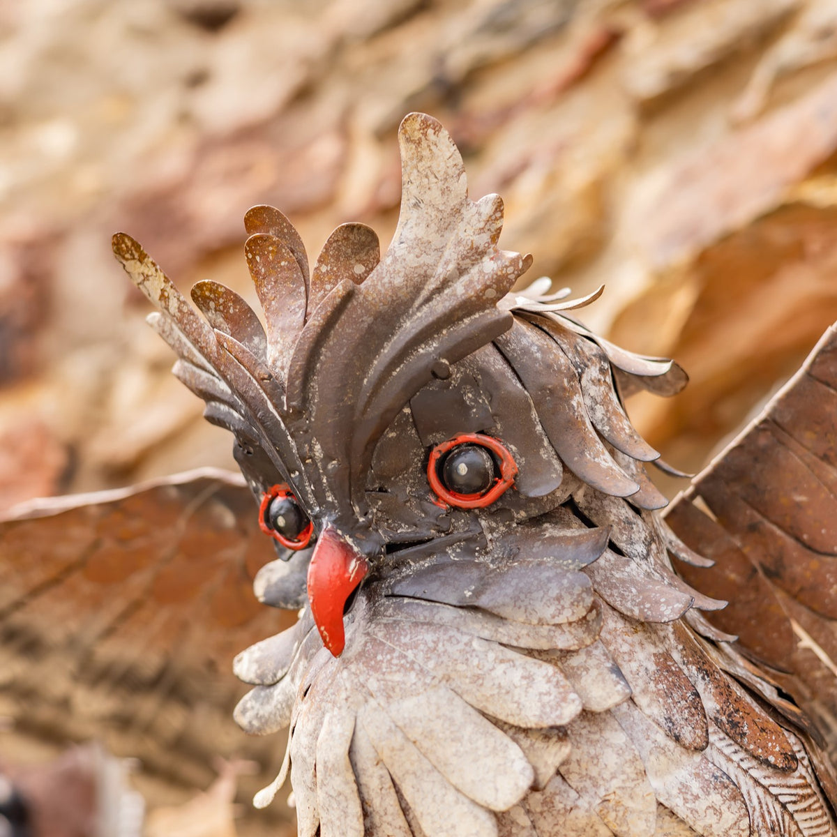 Close view of a metal owl’s layered feathers, bright orange eyes, and red beak against a blurred stone background.