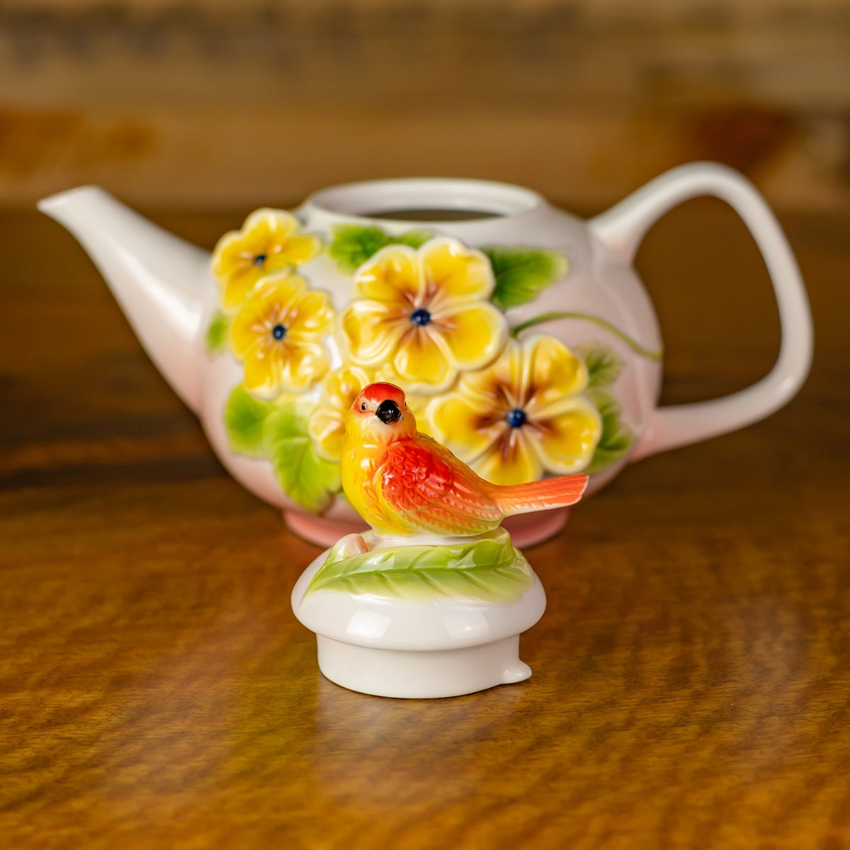 White teapot with raised yellow flowers and a colorful bird ornament on its lid, displayed on a wooden surface.