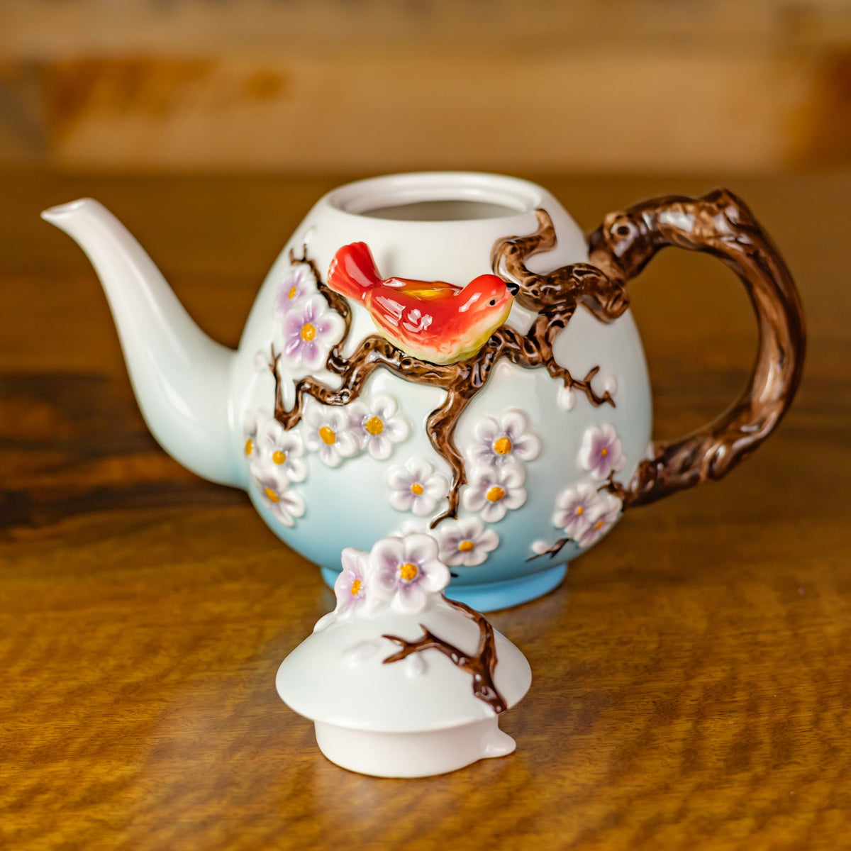 White teapot with raised blossoms, a red bird, and a branch-like handle, with its flower-topped lid set in front.
